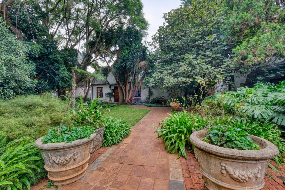 A landscaped garden featuring a paved brick pathway winding through lush greenery, including large decorative stone planters filled with green plants, ferns, and flowering foliage. The pathway is bord